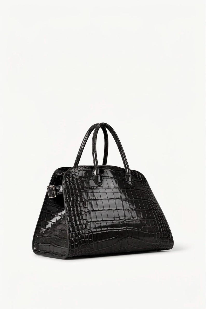 Puede incluir: Un bolso negro estructurado con textura brillante de piel de cocodrilo. Cuenta con dos asas superiores redondeadas y un cierre de hebilla plateada. La forma trapezoidal y el acabado brillante le dan un aspecto lujoso.