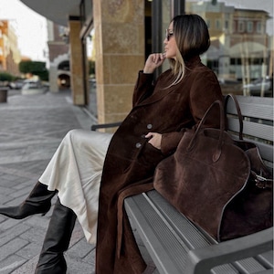 May include: A woman wearing a long brown suede coat, black boots, and a cream-colored dress, seated on a bench. A large brown suede tote bag rests beside her. The scene is outdoors, with a building in the background.