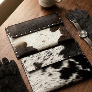May include: Three cowhide leather clutch wallets with silver rivet detailing. The wallets are black and white, and brown and white. A pair of black gloves, a watch with a brown leather band, and a cup of coffee are also on the wooden surface.