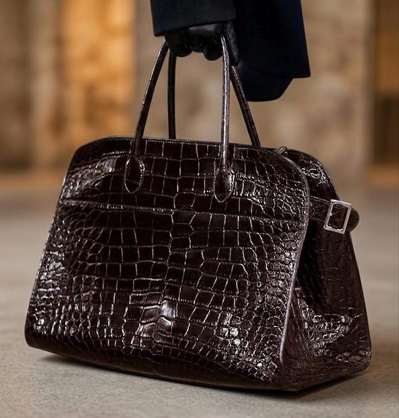 Puede incluir: Un bolso de mano marr&oacute;n oscuro, con textura de cocodrilo, de forma rectangular y dos asas superiores. El bolso tiene un cierre de hebilla plateada en el lateral. El bolso lo sostiene una persona con guantes negros.