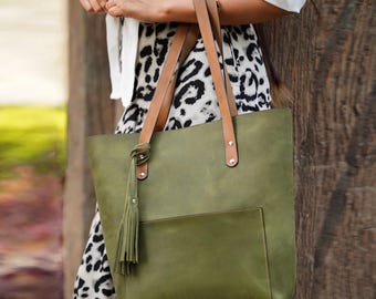 Pode incluir: Bolsa tote verde-oliva com alças de couro marrom e um bolso frontal. A bolsa apresenta uma borla decorativa e detalhes de rebites. A bolsa é feita de um material liso.