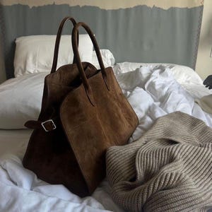 May include: A brown suede tote bag with two handles and a silver buckle closure. A beige knit sweater is next to the bag. The items are on a bed with white sheets and a gray and beige headboard.
