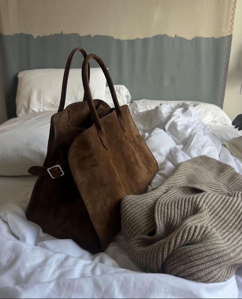 May include: A brown suede tote bag with two handles and a silver buckle closure. A beige knit sweater is next to the bag. The items are on a bed with white sheets and a gray and beige headboard.