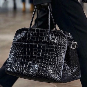 Puede incluir: Un bolso de mano de cuero negro texturizado con un patr&oacute;n de cocodrilo. El bolso tiene dos asas superiores y un cierre de hebilla plateada. La persona lleva ropa negra.