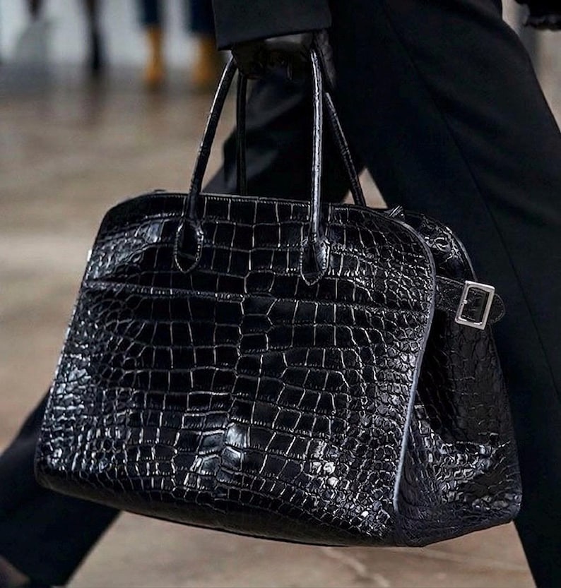 Puede incluir: Un bolso de mano de cuero negro texturizado con un patr&oacute;n de cocodrilo. El bolso tiene dos asas superiores y un cierre de hebilla plateada. La persona lleva ropa negra.