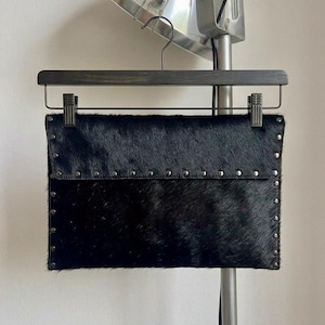 May include: A black clutch made of textured material, possibly animal hide, with a flap closure. The clutch is adorned with small, silver-colored studs along the edges and flap. It hangs from a wooden hanger.