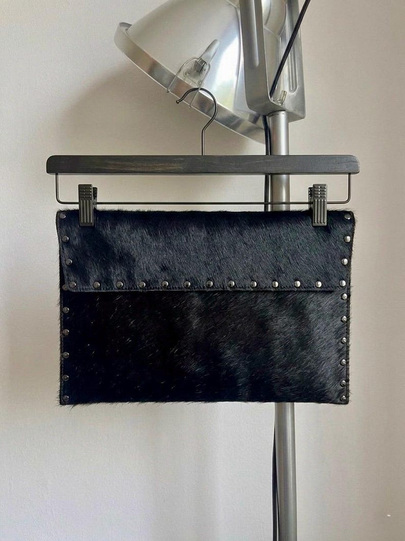 May include: A black clutch made of textured material, possibly animal hide, with a flap closure. The clutch is adorned with small, silver-colored studs along the edges and flap. It hangs from a wooden hanger.