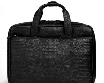 May include: Black briefcase with a textured, crocodile-like pattern. The bag features a top handle, a front zipper pocket, and a main compartment with a zipper closure. The bag is rectangular and appears to be made of leather.