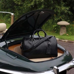 May include: A black leather holdall with a textured finish rests in the open boot of a classic green car. The bag has a shoulder strap and a small tag. The car is parked outside.