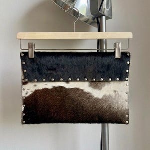 May include: A rectangular clutch made of cowhide, featuring a two-tone design with black and brown fur separated by a white section. Silver-colored studs line the edges. The clutch is suspended from a wooden hanger.