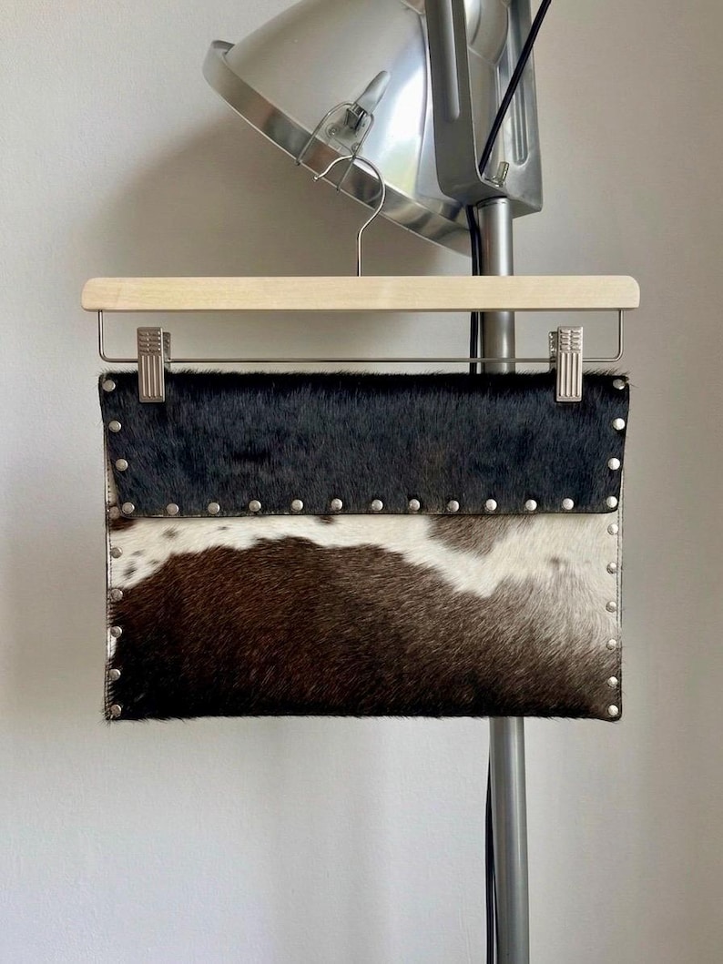 May include: A rectangular clutch made of cowhide, featuring a two-tone design with black and brown fur separated by a white section. Silver-colored studs line the edges. The clutch is suspended from a wooden hanger.
