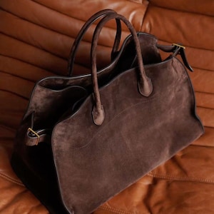 May include: A brown suede tote bag with two handles and a gold buckle closure. The bag has a spacious interior and a classic design, perfect for everyday use. The bag is made of a soft, luxurious suede material.