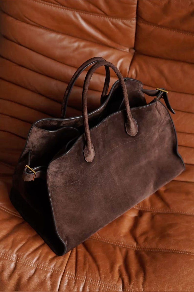 May include: A brown suede tote bag with two handles and a gold buckle closure. The bag has a spacious interior and a classic design, perfect for everyday use. The bag is made of a soft, luxurious suede material.