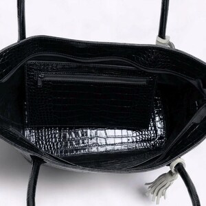 May include: Black tote bag with a crocodile skin texture. The bag has a zippered interior pocket and a matching exterior texture. The handles are black with a light gray accent and a tassel.