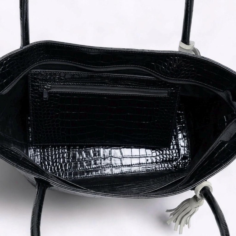 May include: Black tote bag with a crocodile skin texture. The bag has a zippered interior pocket and a matching exterior texture. The handles are black with a light gray accent and a tassel.