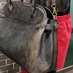 May include: A distressed leather handbag with a top handle and a detachable shoulder strap. The bag is dark brown with lighter brown accents and gold-colored hardware. The bag has a classic, structured design.
