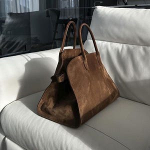 May include: A brown suede handbag with two handles and a buckle closure. The bag is trapezoidal in shape and sits on a white leather couch. The bag has a soft, textured appearance.