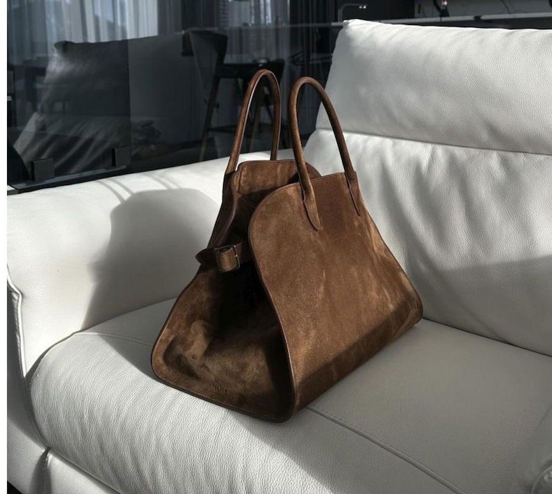 May include: A brown suede handbag with two handles and a buckle closure. The bag is trapezoidal in shape and sits on a white leather couch. The bag has a soft, textured appearance.