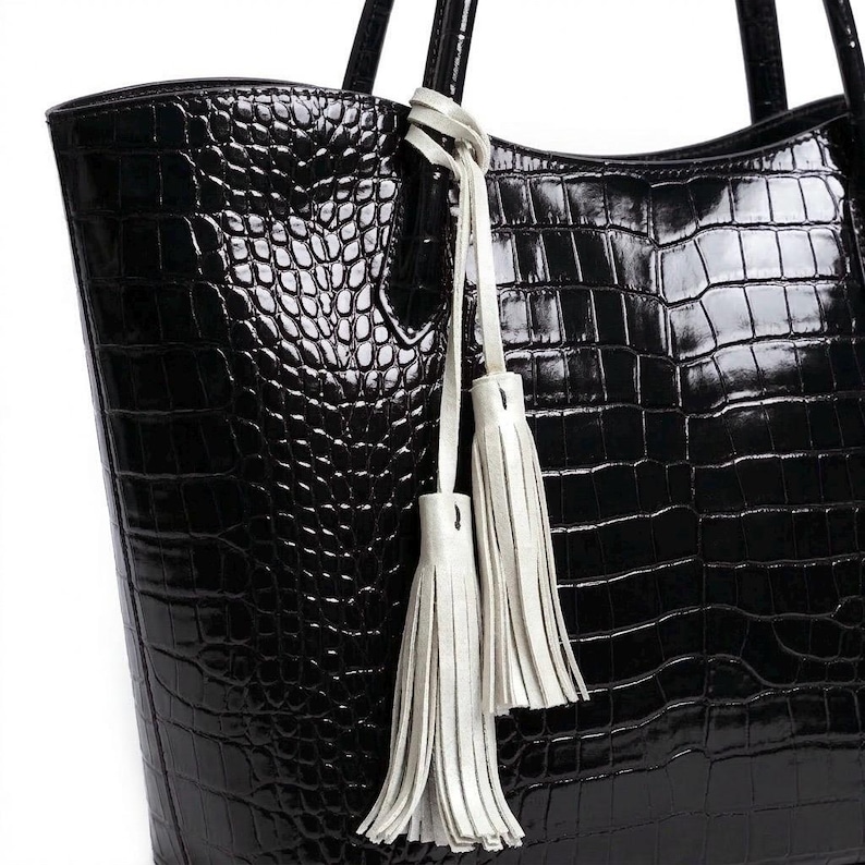 May include: Black tote bag with a crocodile skin texture. The bag has two handles and is adorned with two white leather tassels hanging from the handle. The bag is made of a shiny, patent leather material.