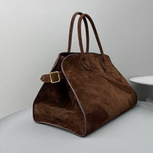 Puede incluir: Un bolso de mano de ante marrón con dos asas superiores y un cierre de hebilla rectangular dorado. El bolso tiene una forma estructurada con una parte superior redondeada y un ribete marrón oscuro.