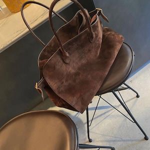 May include: A large, brown suede tote bag with two rounded handles and a rectangular shape. The bag is resting on a brown leather chair with a black metal frame. The chair has a rounded seat. A second chair is in the foreground.