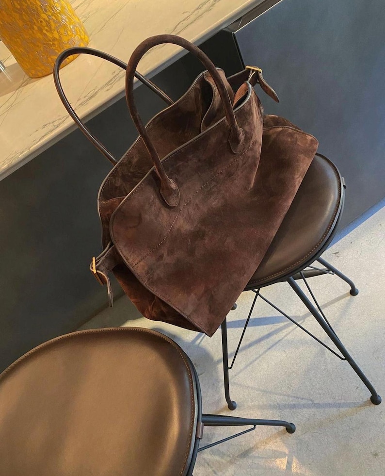 May include: A large, brown suede tote bag with two rounded handles and a rectangular shape. The bag is resting on a brown leather chair with a black metal frame. The chair has a rounded seat. A second chair is in the foreground.