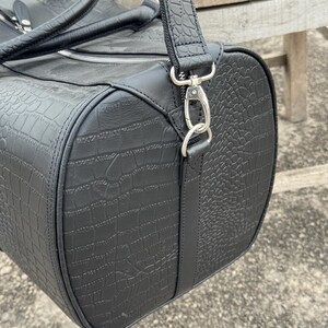 Op de afbeelding: Zwarte duffeltas met een krokodillenleertextuur. De tas heeft een cilindrische vorm met een ritssluiting en een afneembare schouderriem. De tas heeft een handvat aan de bovenkant en zilverkleurige metalen hardware.