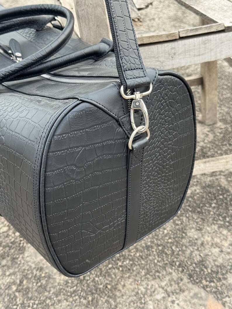 Op de afbeelding: Zwarte duffeltas met een krokodillenleertextuur. De tas heeft een cilindrische vorm met een ritssluiting en een afneembare schouderriem. De tas heeft een handvat aan de bovenkant en zilverkleurige metalen hardware.