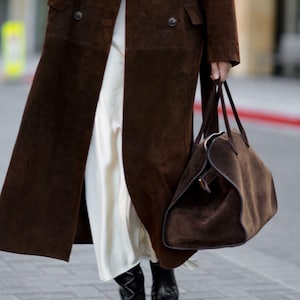 May include: A long, dark brown suede coat with large buttons and a matching suede tote bag. The coat is worn over a white dress, and the outfit is completed with black leather boots. The overall style is classic and elegant.