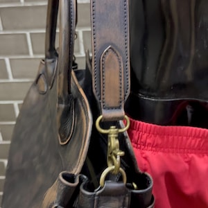 May include: A dark brown leather tote bag with a shoulder strap and a top handle. The bag has brass-colored hardware and a distressed finish, suggesting a vintage or rustic style. The bag is designed for carrying items.