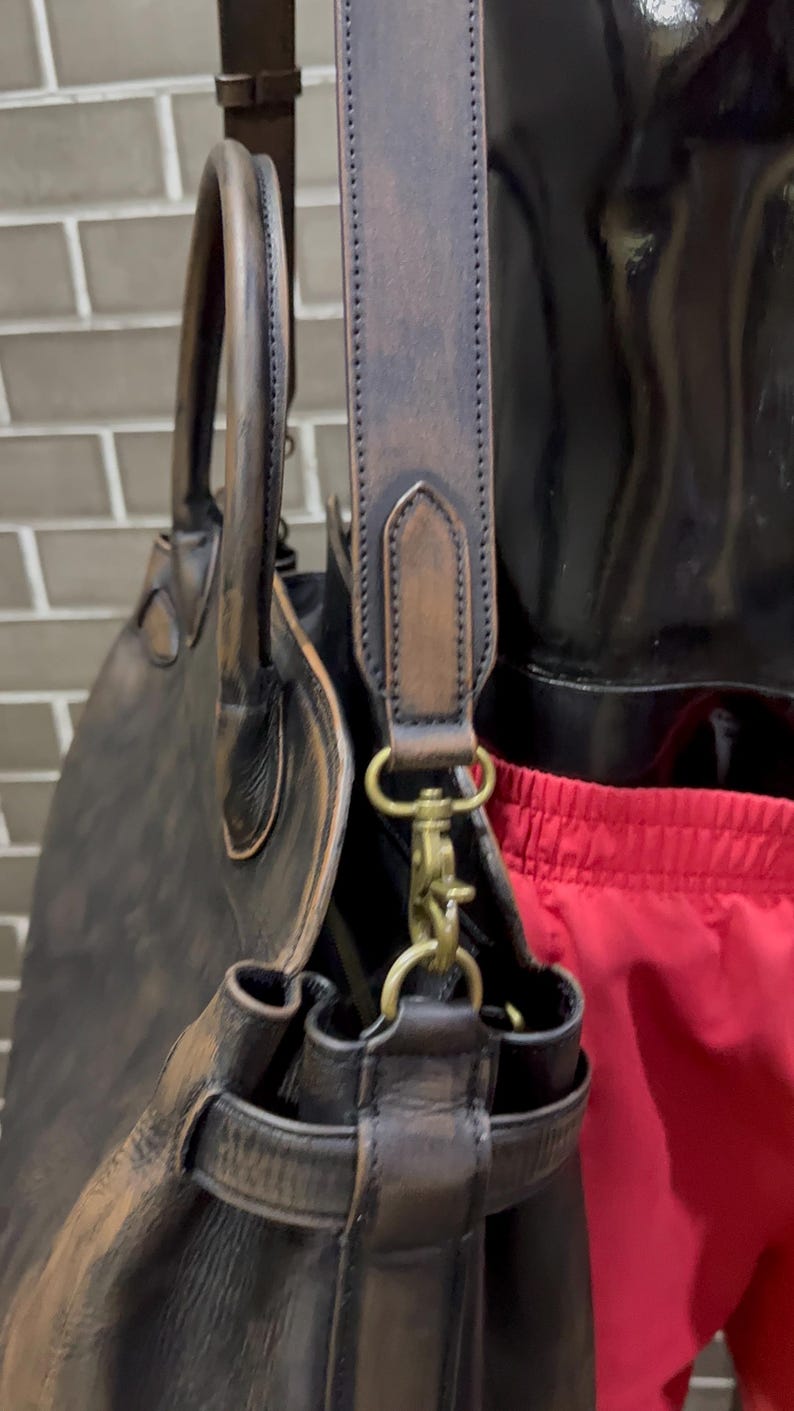 May include: A dark brown leather tote bag with a shoulder strap and a top handle. The bag has brass-colored hardware and a distressed finish, suggesting a vintage or rustic style. The bag is designed for carrying items.