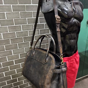 May include: A distressed brown leather satchel bag with a shoulder strap and top handles. The bag has a worn, vintage appearance. The bag is displayed on a black mannequin torso with red shorts.