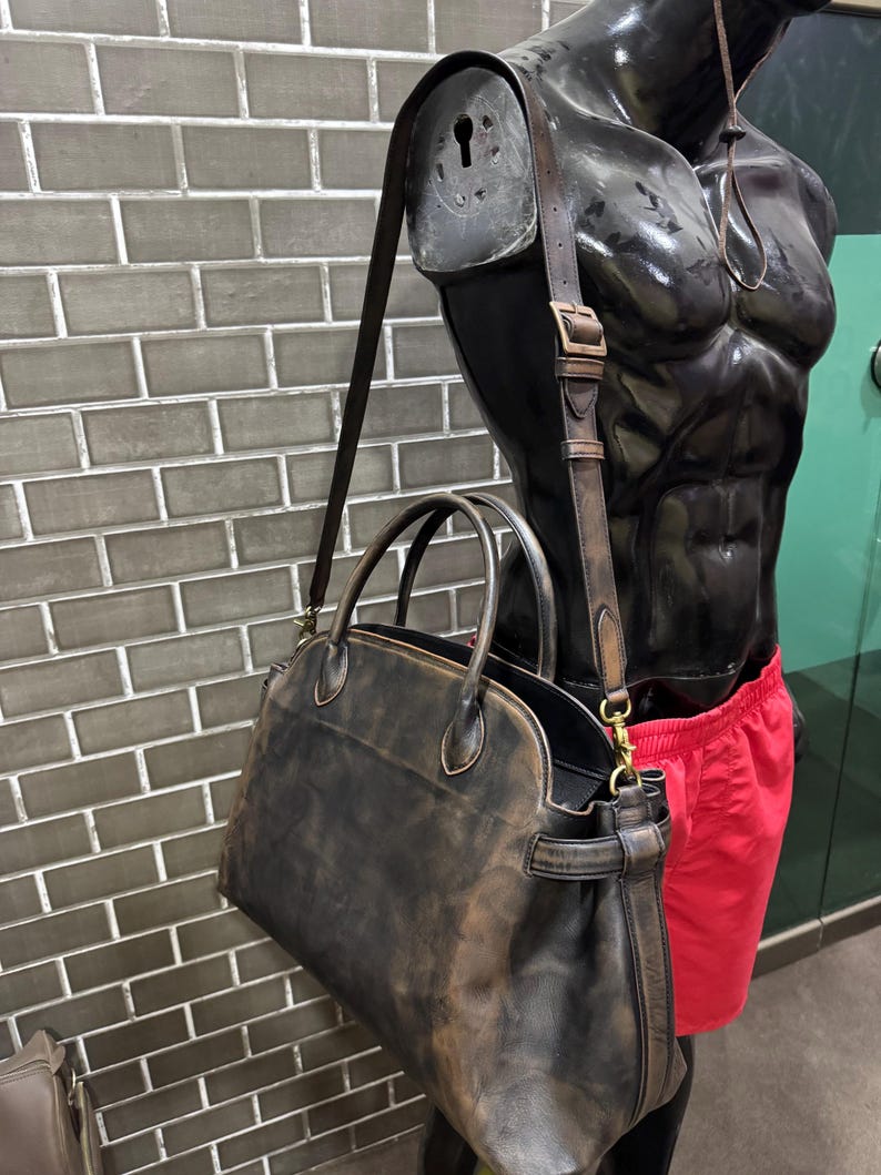 May include: A distressed brown leather satchel bag with a shoulder strap and top handles. The bag has a worn, vintage appearance. The bag is displayed on a black mannequin torso with red shorts.