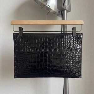 May include: A black clutch with a crocodile skin texture, held by a wooden hanger. The rectangular bag has a row of small silver studs along the top. The bag is hanging in front of a silver lamp.