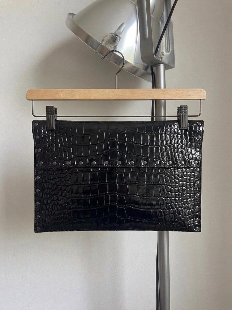 May include: A black clutch with a crocodile skin texture, held by a wooden hanger. The rectangular bag has a row of small silver studs along the top. The bag is hanging in front of a silver lamp.