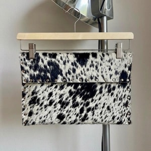 May include: A black and white cowhide clutch with silver-tone stud accents. The rectangular bag is suspended from a wooden hanger. The spotted pattern is a striking feature of this accessory.