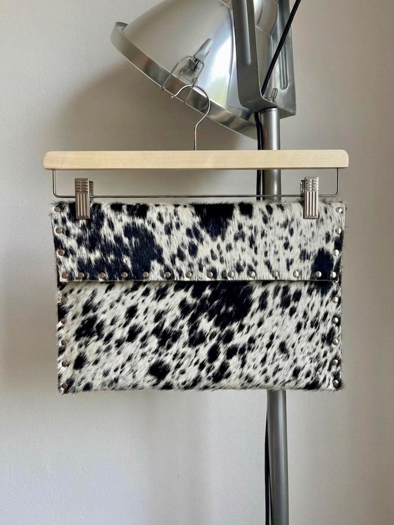 May include: A black and white cowhide clutch with silver-tone stud accents. The rectangular bag is suspended from a wooden hanger. The spotted pattern is a striking feature of this accessory.