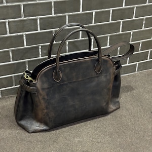 May include: A dark brown leather handbag with two top handles and a detachable shoulder strap. The bag has a rounded shape and a distressed finish, giving it a vintage look. The bag is sitting on a gray surface.