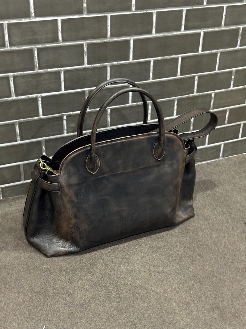 May include: A dark brown leather handbag with two top handles and a detachable shoulder strap. The bag has a rounded shape and a distressed finish, giving it a vintage look. The bag is sitting on a gray surface.