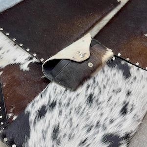 May include: A cowhide wallet with a flap closure, featuring a mix of brown, black, and white patterns. The wallet is set on a patchwork of cowhide pieces, some with silver stud accents.