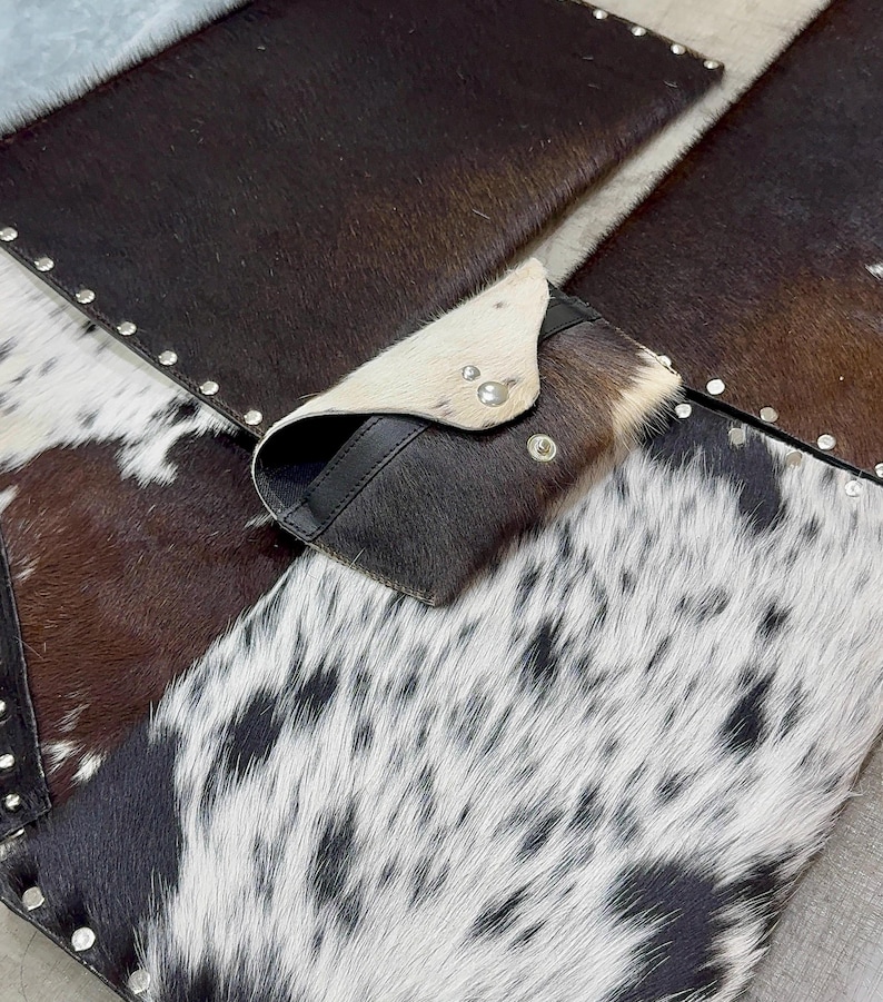 May include: A cowhide wallet with a flap closure, featuring a mix of brown, black, and white patterns. The wallet is set on a patchwork of cowhide pieces, some with silver stud accents.