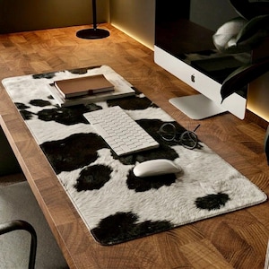 May include: A desk mat with a cowhide pattern in black and white. A white keyboard, mouse, and round-framed glasses sit on the mat. A brown notebook and a computer monitor are also visible.