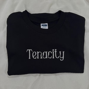 May include: Black crew neck sweatshirt with the word "Tenacity" embroidered in white script across the chest. The sweatshirt is folded neatly, and the fabric appears to be a soft material. The tag reads "H&U Regular Fit".