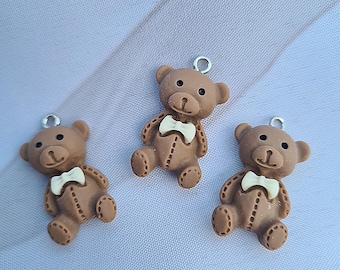 Op de afbeelding: Drie bruine teddybeerbedels met witte vlinderdassen en zilveren springringen. De beren hebben knoopdetails en zitten rechtop. De achtergrond is een lichtblauwe stof.