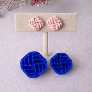 Könnte beinhalten: Ein Paar rosa Ohrstecker im Webstil, präsentiert auf einem beigen Ständer. Darunter sind zwei blaue Formen desselben Designs zu sehen. Die Ohrringe und Formen haben eine quadratische Form mit einem ineinander verwobenen Muster.