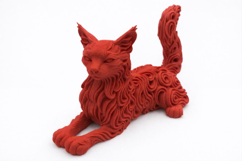 Op de afbeelding: Een rode kattenbeeldhouwwerk in een ontspannen pose. De vacht van de kat is gedetailleerd met wervelende patronen en de staart is gekruld. Het beeldhouwwerk staat op een witte achtergrond, wat de vorm en textuur van de kat benadrukt. Een decoratief item.