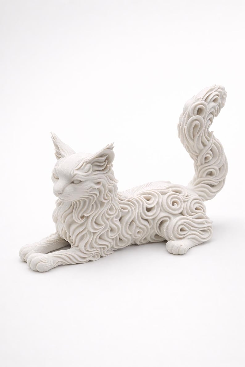 Op de afbeelding: Een witte kattenfiguur in een liggende houding. De vacht van de kat is gedetailleerd met een wervelend, gestructureerd patroon. De sculptuur heeft puntige oren en een gekrulde staart, wat een decoratief en artistiek stuk cre&euml;ert.