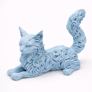 Op de afbeelding: Een lichtblauwe kattenfiguur in rustpositie. De vacht van de kat is gedetailleerd met een wervelend patroon. De figuur heeft puntige oren en een lange, gekrulde staart. De kat ligt op een witte ondergrond.