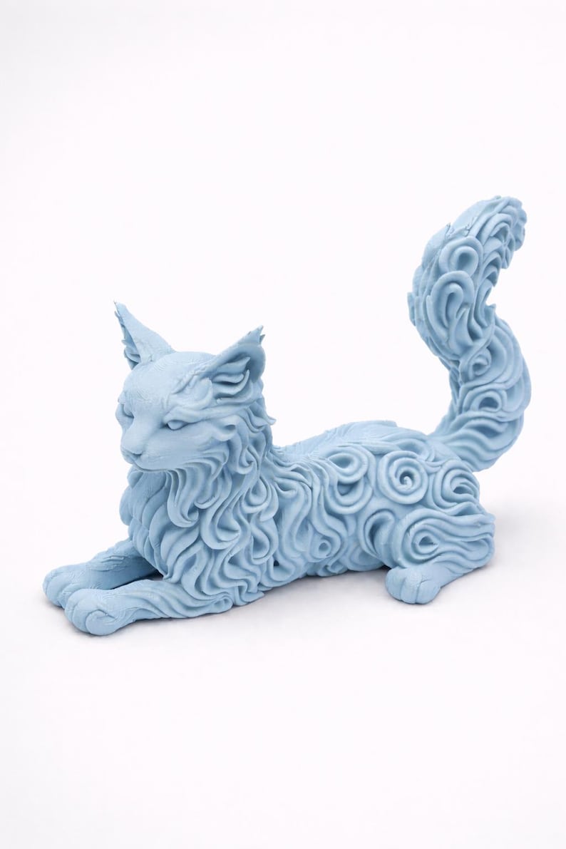 Op de afbeelding: Een lichtblauwe kattenfiguur in rustpositie. De vacht van de kat is gedetailleerd met een wervelend patroon. De figuur heeft puntige oren en een lange, gekrulde staart. De kat ligt op een witte ondergrond.