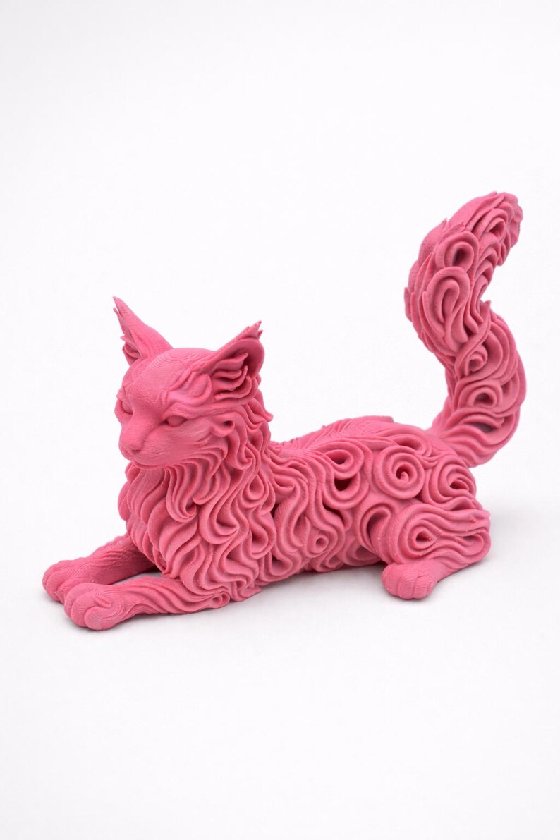 Op de afbeelding: Een roze kattenbeeld in een liggende houding. De vacht van de kat is gedetailleerd met een wervelend patroon. De staart is omhoog gekruld. De sculptuur staat op een witte achtergrond.